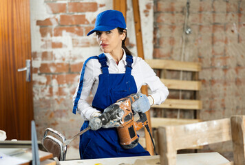 Skilled female builder in blue overalls confidently holding pneumatic jackhammer while standing inside house under construction