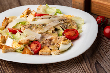 A Caesar salad with grilled chicken breasts, lettuce, croutons, eggs, cherry tomatoes, dressed with Parmesan cheese and sauce. On wooden background.