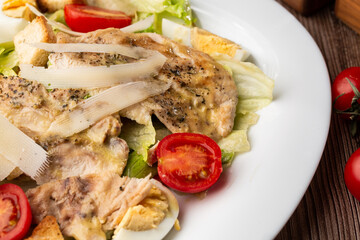A Caesar salad with grilled chicken breasts, lettuce, croutons, eggs, cherry tomatoes, dressed with Parmesan cheese and sauce. On wooden background.