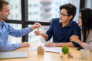 Caucasian male real estate agent giving house key to happy couple after sign contract in office. Asian couple receiving new house or apartment key from broker. New house and relocation concept.