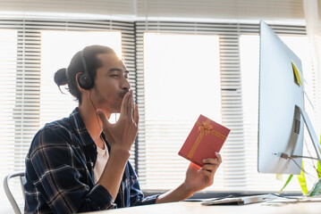 Happy young Asian man showing gift box while having video call on computer with his girlfriend or...