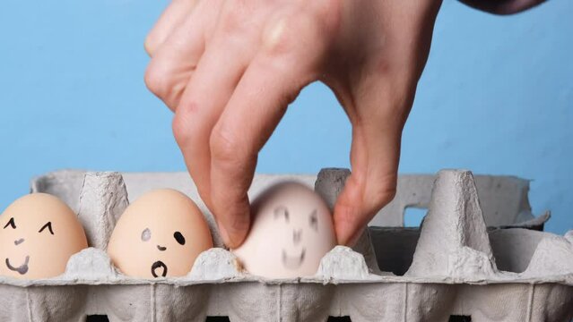 Hand folds eggs with smileys in an egg tray on blue background. Easter concept. Cultural symbol of Easter. Farm eggs with a drawn faces.