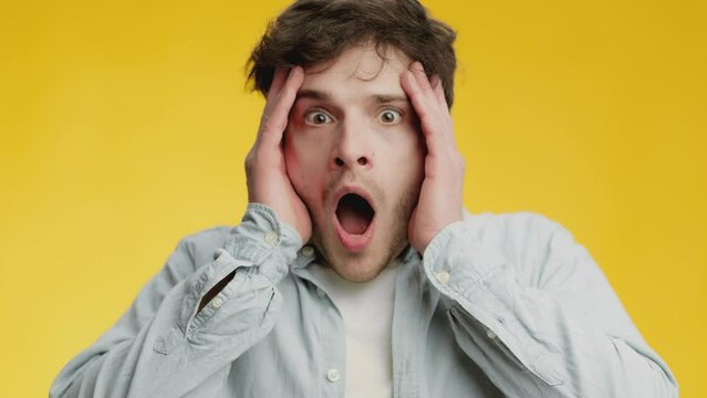 Young man looking to camera attentively and feeling shocked, touching face in amazement with open mouth