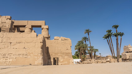 Ruins of the Karnak Temple in Luxor. Dilapidated stone walls with carved drawings, hieroglyphs, columns are visible. Tall palm trees against a clear blue sky Egypt