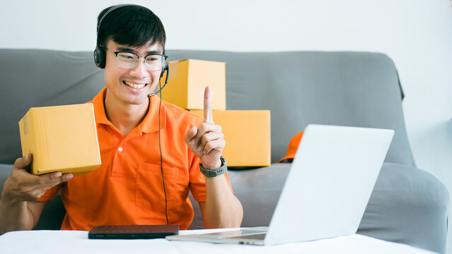 Handsome Asian Thai Male Employee Is Contacting Customer Via Laptop Webcam To Sell In The Packaging Box.Online Order Confirmation Working At Home In Orange Uniforms.Ecommerce Internet Work From Home.