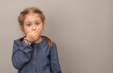 Little girl cover her mouth with hand over grey background. Shocked and surprised child in...