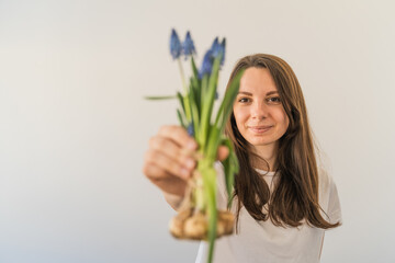 Caucasian woman in white t-shirt holds a bunch of blue muscari flowers with bulbs in her hand. White background.