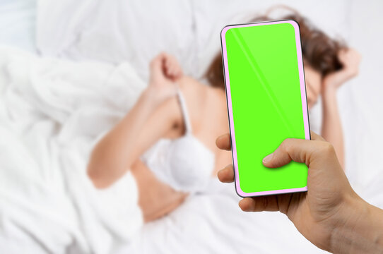 Male hand using smartphone with blank green screen to take photo of sensual woman in bed