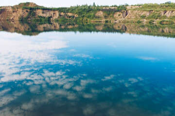 Picturesque blue lake summer landscape for outdoors vacation. Blue sky and clouds reflection in clear pond water with lush greenery forest in the background.