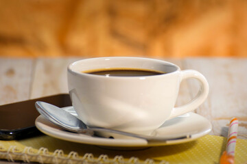 A hot cup of coffee, a notebook, a pen and a smartphone on the table.