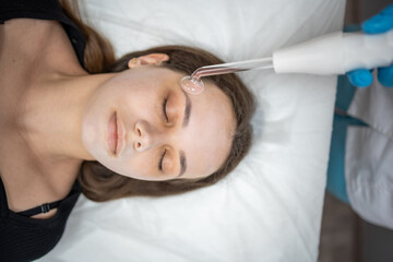 Obraz na płótnie Canvas Beautician uses the darsonval device on the client's face. Professional skin care in the salon. The concept of cosmetology and rejuvenation.