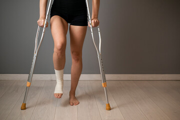Front view close up of a disabled woman walking with crutches and sprained bandaged ankle at home
