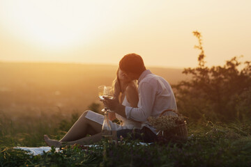 Boyfriend and girlfriend holding wine glasses with beverage and sitting on blanket at summer picnic at sunset. They want to kiss. 