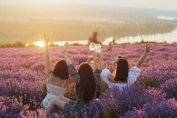 Girlfriends having picnic in the lavender field at sunset. Group of young women sitting on lavender field on summer day. Girlfriends drinking wine on outdoor party.