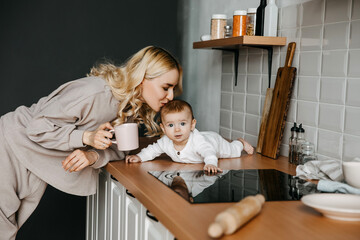 Mother and son at the kitchen at home, wearing pajamas. Woman playing with her baby, kissing his head and drinking coffee in the morning.