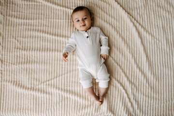 4 months old baby wearing white overall, lying on a soft blanket on bed at home, smiling, top view.