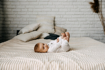 4 months old baby wearing white overall, lying on a soft blanket on bed at home, barefoot.