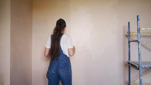 Professional painter female paints the wall with roller and tray on construction site. Young woman in overalls and cap doing home decoration and renovetion. Worker makes DIY repair work in the room.