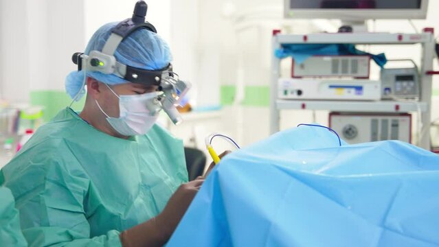 Busy male doctor with device glasses on head operating the patient. Specialist holds an instrument and takes a large syringe.