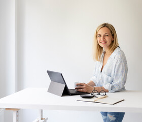 pretty young blonde business woman stands by her ergonomic height adjustable work desk and works on tablet from home in her home office