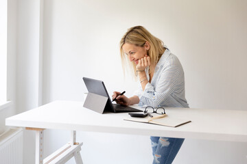 pretty young blonde business woman stands by her ergonomic height adjustable work desk and works on tablet from home in her home office