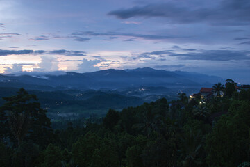 Fototapeta na wymiar Indonesian tender landscape - sunrise with fresh morning sky, light pink clouds, sunbeams over hazy blue mountains, green valley, tropical forest, houses with yellow lights on Bali, Munduk village.