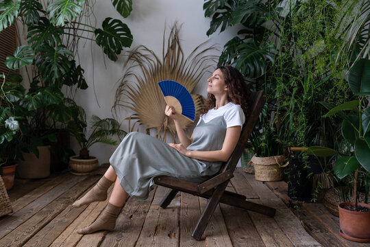 Stress relief after work from home. Relaxed young lady spend time in cozy indoor garden with monstera plant sitting with closed eyes and blowing fan. Love for plants, wellness and wellbeing concept