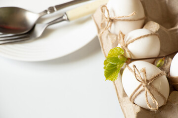 Knife and fork in a plate on a white table and next to a tray with chicken eggs decorated with a rope with a bow, easter holiday
