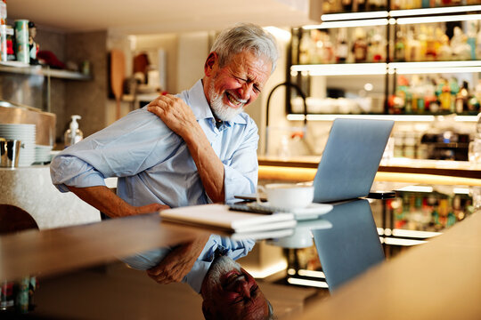 A senior man sits in a coffee shop and has back pain.