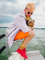 handsome blond young person wear raincoat sitting on a sailing yacht and hold his small dog...
