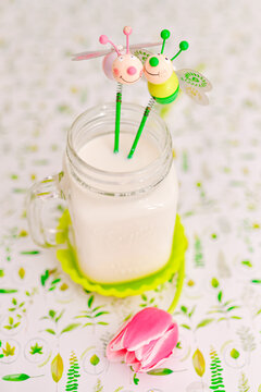 A pot of milk decorated by two smiling bees; all placed on a spring decorated tablecloth with yellow and green leaves details and a pink tulip lying around.