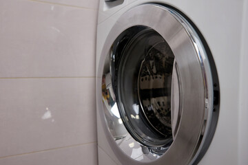 A modern washing machine with a metal door standing near a tiled wall in the room, a place for text. Bathroom interior