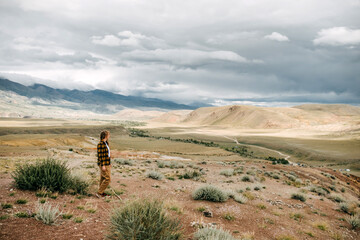 A young woman with blond hair stands in a chasing valley in cloudy weather and looks into the distance. Travel content. Mars field, Altai