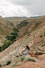 A young brunette woman with curly hair sits on a cliff overlooking a mountain valley. Altai, Mars Valley