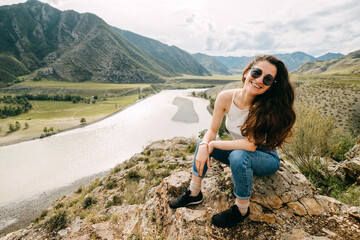 A young brunette Armenian woman with curly hair in black sunglasses sits and enjoys the mountain view and the river. Altai. Confluence of Chuya and Katun