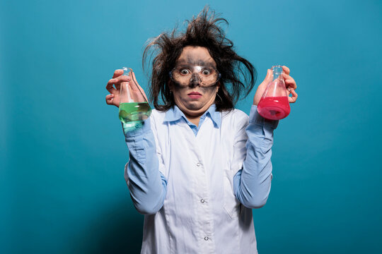 Mad chemist with dirty face and messy hair having flasks filled with unknown chemical substances. Surprised crazy chemistry specialist holding glass beakers filled with dangerous liquid compounds.