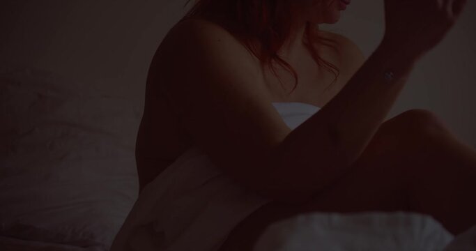 Naked red-haired woman covering herself with white sheets in a dimly lit dark environment