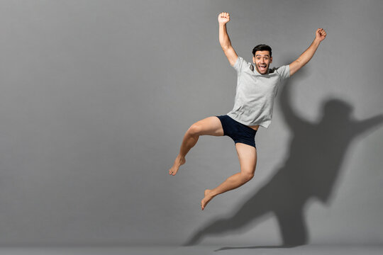 Surprised caucasian man in sleepwear jumping and raising fists on gray isolated background in studio with copy space