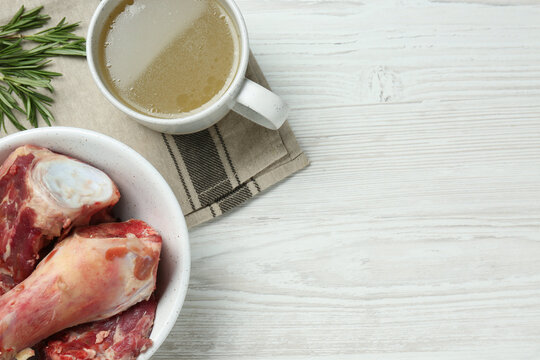 Mug with delicious bone broth and ingredients on white wooden table, flat lay. Space for text