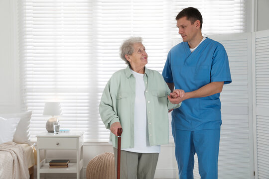 Caregiver assisting senior woman with walking cane indoors. Home health care service