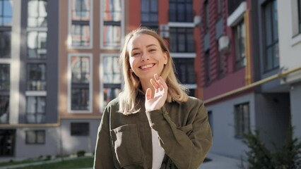 Cute blonde woman walking on the urban street looking at camera. Portrait of beautiful happy girl with healthy white teeth standing in city near beautiful colored buildings. Positive emotion. Freedom.