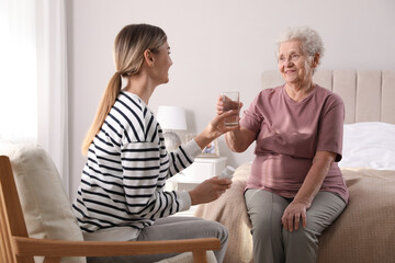 Young caregiver giving water to senior woman in bedroom. Home care service