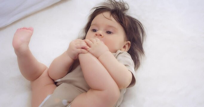 Cute girl in a beige bodysuit lies on her back on a white bed in the nursery, plays and chewing foot. The concept of love, parenthood, childhood, life, motherhood