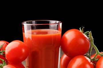 A glass cup with tomato juice, next to it lies a branch of tomatoes. On a wooden table there is a glass of tomato juice and tomatoes lie on a black background.