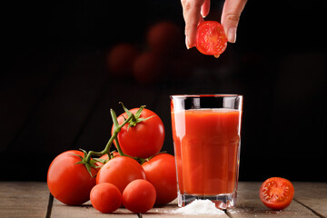 A glass cup with tomato juice, next to it lies a branch of tomatoes. On a wooden table there is a glass of tomato juice and tomatoes lie on a black background.Hand holding a tomato over a glass of jui