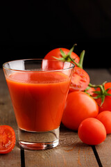 A glass cup with tomato juice, next to it lies a branch of tomatoes. On a wooden table there is a glass of tomato juice and tomatoes lie on a black background.