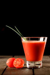 A glass cup with tomato juice, next to it lies a branch of tomatoes. On a wooden table there is a glass of tomato juice and tomatoes lie on a black background.