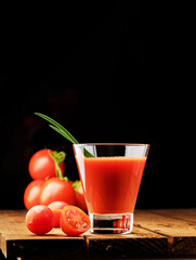 A glass cup with tomato juice, next to it lies a branch of tomatoes. On a wooden table there is a glass of tomato juice and tomatoes lie on a black background.