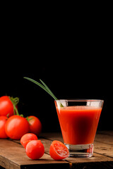 A glass cup with tomato juice, next to it lies a branch of tomatoes. On a wooden table there is a glass of tomato juice and tomatoes lie on a black background.