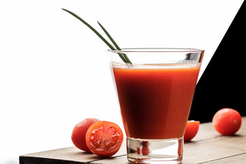 A glass cup with tomato juice, next to it lies a branch of tomatoes. On a wooden table there is a glass of tomato juice and tomatoes lie on a black background.